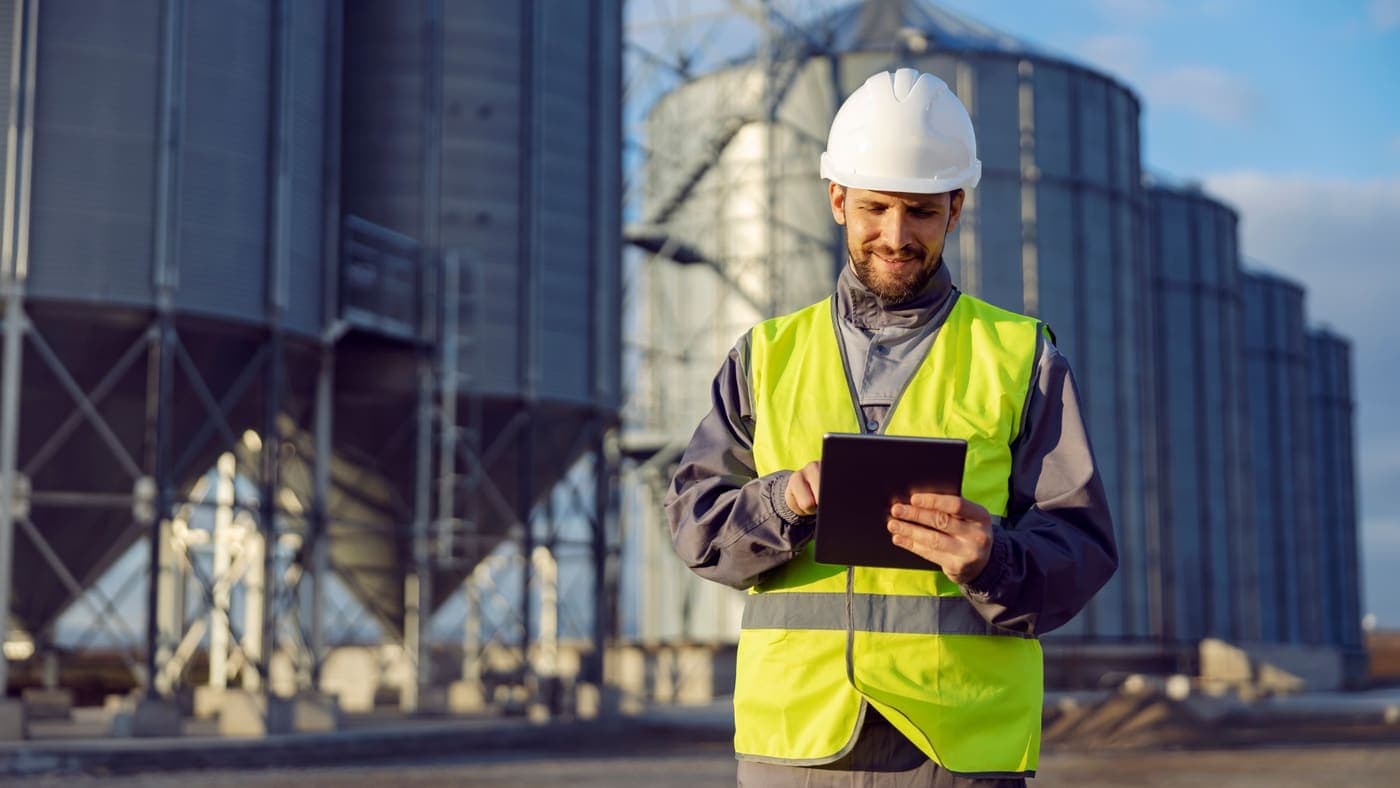 A worker in a hard hat and a high-visibility vest using a tablet in front of grain silos.